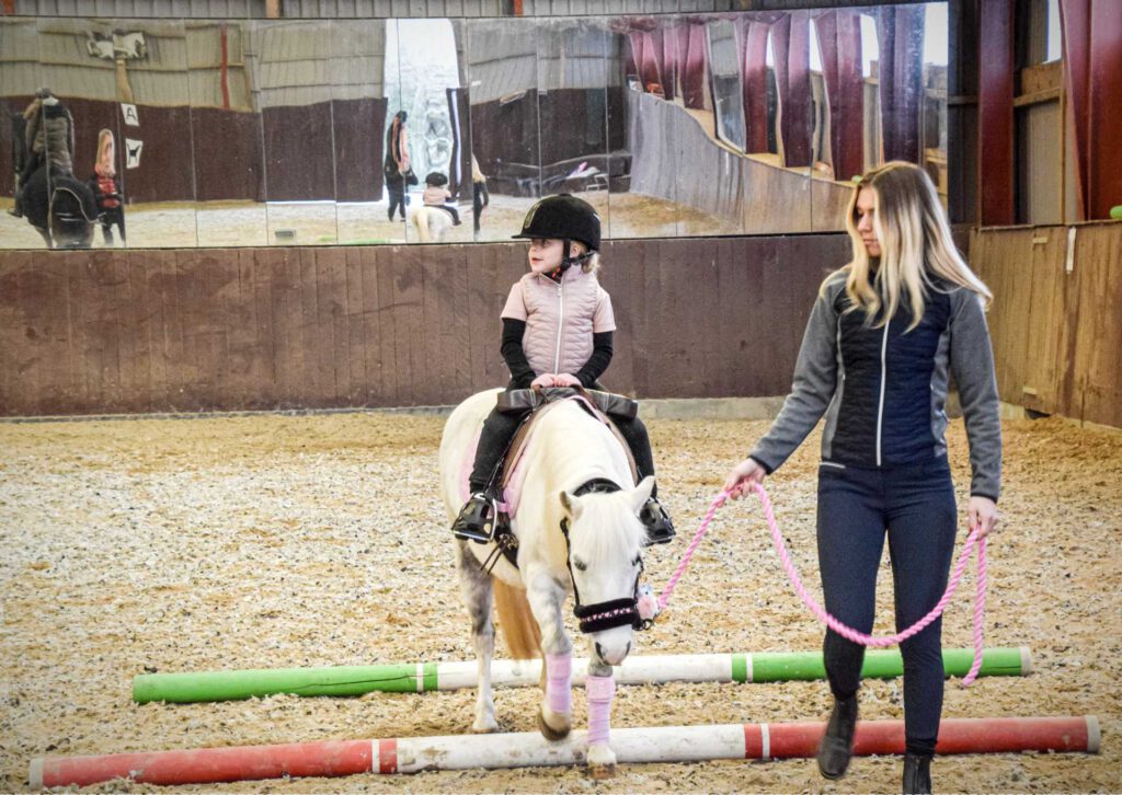 Et barn træner sin hest i ridehus med hjælp fra en forælder